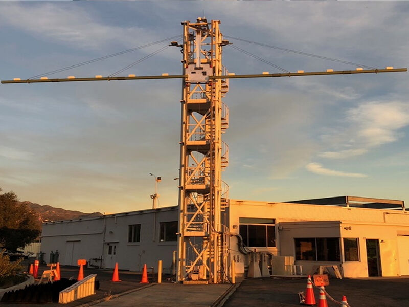 Image shows a building in the late afternoon sun and open sky with light clouds. A large, narrow, and tall tower structure with stairs is in the center in front of the building. The antenna support support is deployed off both sides of the tower near the top. There are some small wire supports attaching the long fixture to the top of the tower.  (Coutesy NASA/JPL-CalTech)